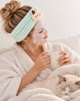 Woman wearing a fluffy mint green cat spa headband and face mask while relaxing with her cat.