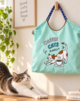 Mint green Coffee Cats tote bag hanging on a wall above a stretching cat in a bright home.