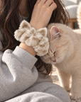 A person wears a soft, fluffy cream cat ear scrunchie on their wrist. A cream-colored cat gently rubs its head against the scrunchie.