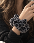 A close-up of a person wearing a fluffy leopard-print hair scrunchie in black and gray, holding an iced coffee. The person is wearing a black ribbed knit sweater.