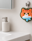 Orange cat-face-shaped hand towel with a light blue scarf and green trim hanging from a wall hook beside a bathroom sink.