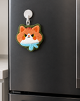 Orange cat-face-shaped hand towel with a light blue scarf and green trim, hanging from a magnetic hook on a refrigerator door.