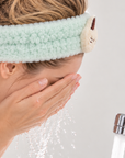 Woman wearing a fluffy mint green cat spa headband while washing her face.