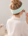Woman wearing a fluffy mint green cat spa headband while applying makeup with a cat beside her.