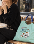 Mint green Coffee Cats tote bag displayed on a chair beside a woman enjoying coffee at an outdoor cafe.