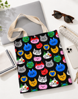 A colorful cat face tote bag alongside everyday essentials including a laptop, sunglasses, smartphone, pencil case, and keys.