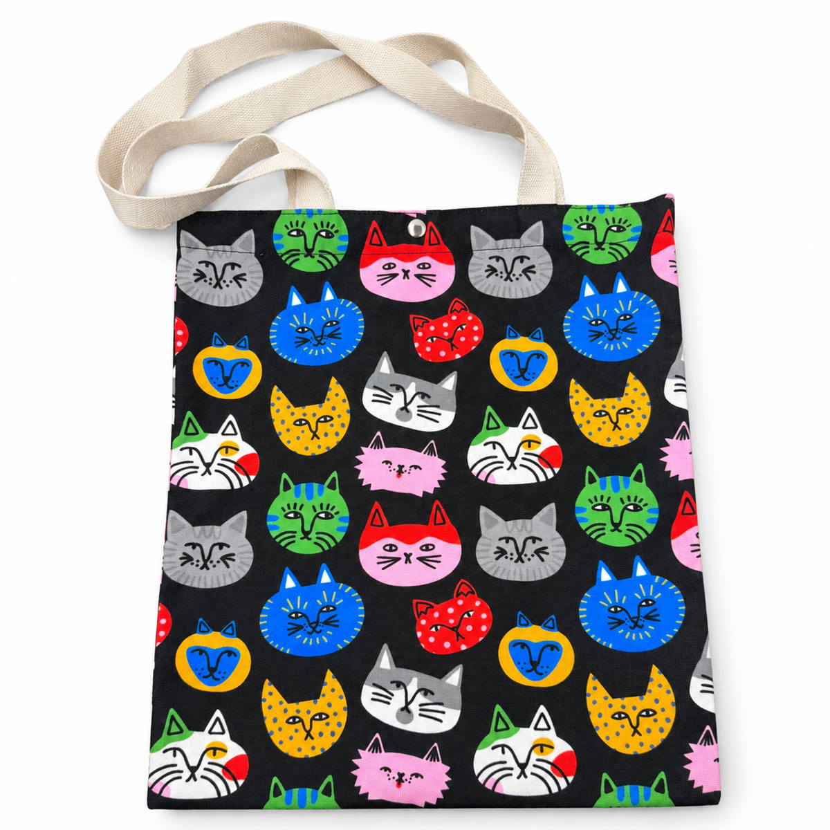 A tote bag with a black background and a colorful repeating pattern of cartoon cat heads in various colors, featuring natural beige shoulder straps.