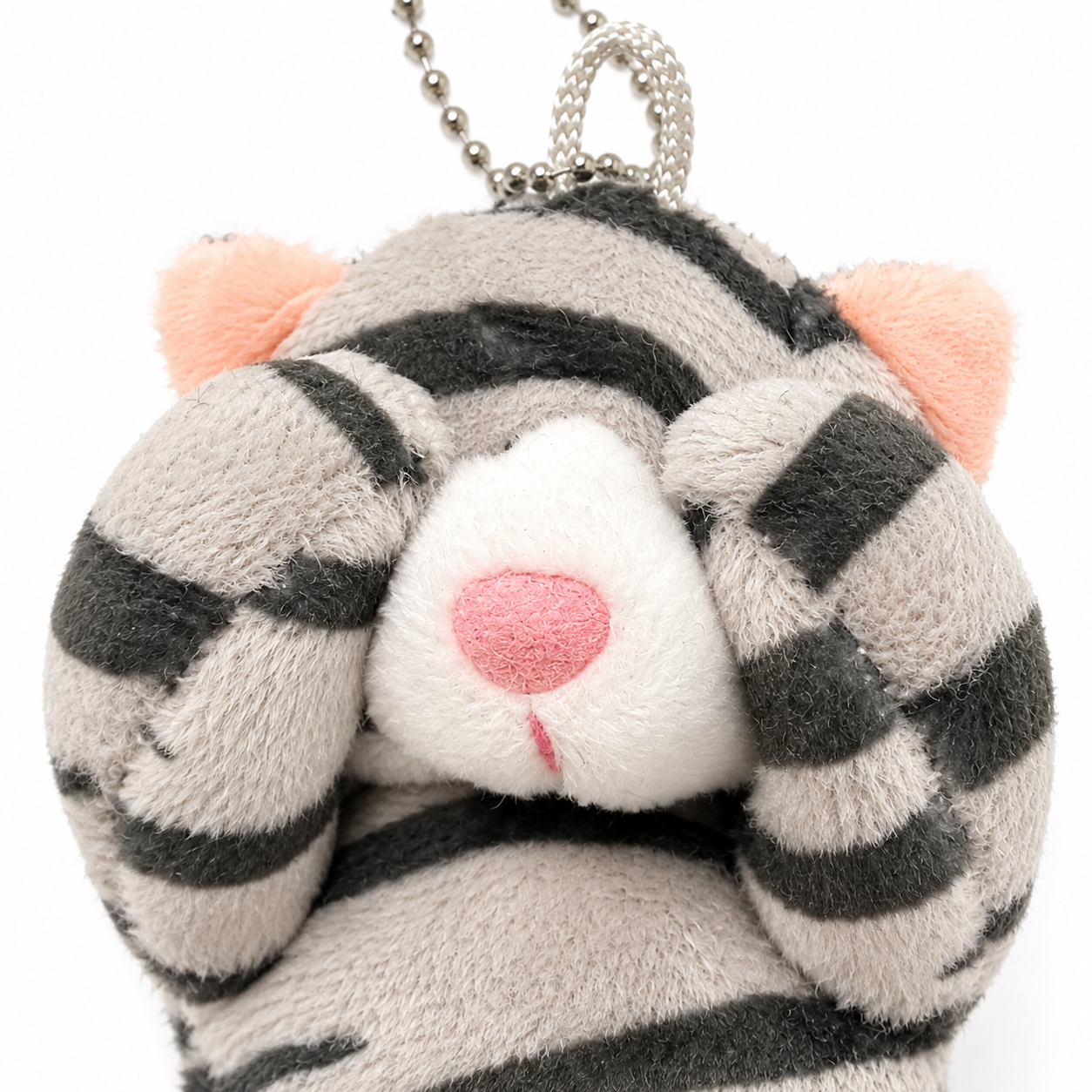 Close-up of a gray tabby cat plush charm with its paws covering its eyes, highlighting its soft fabric texture, white muzzle, pink inner ears, and pink nose.