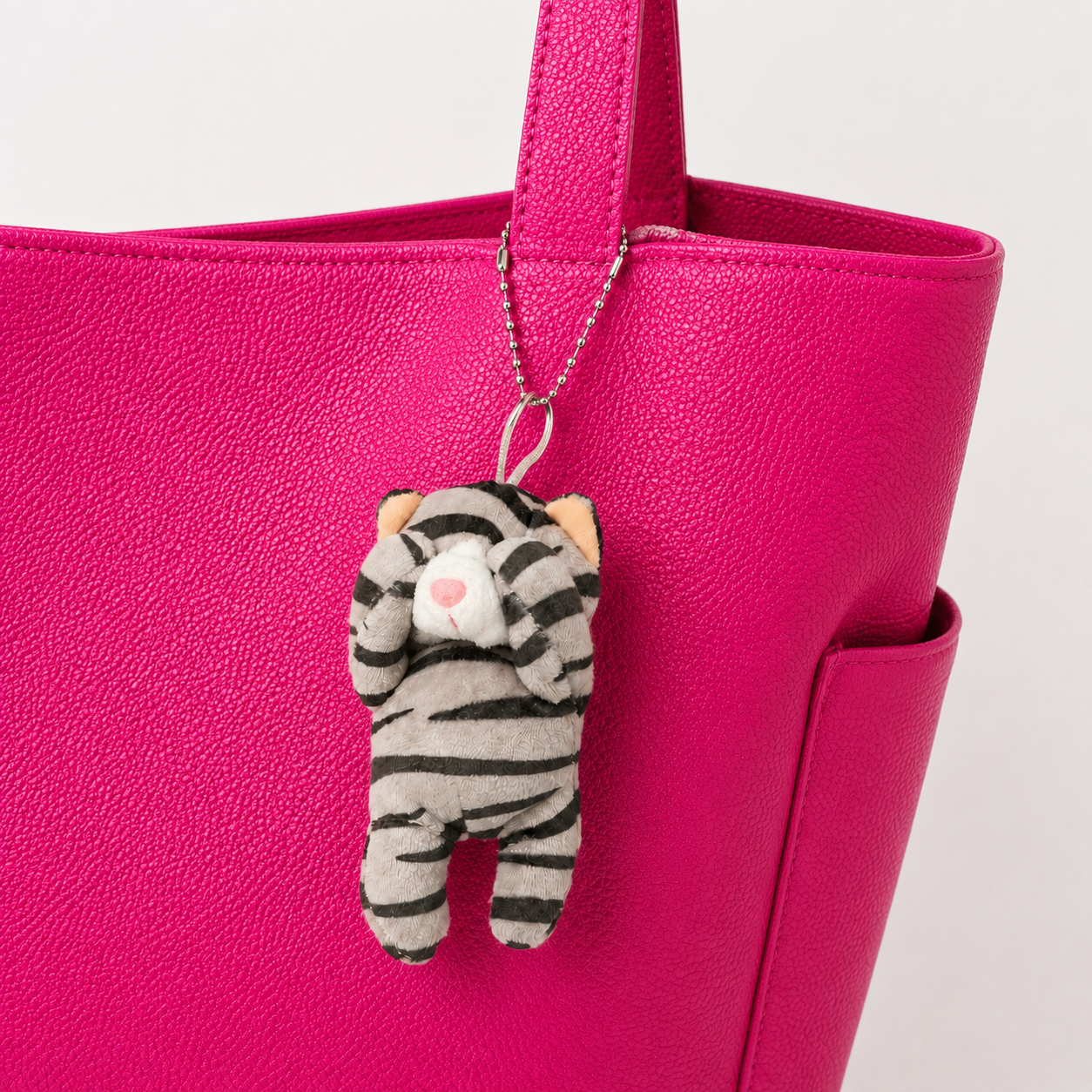 A gray tabby cat plush charm with its paws covering its eyes, hanging from the base of the handle of a pink leather bag by a silver ball chain.