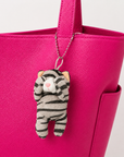 A gray tabby cat plush charm with its paws covering its eyes, hanging from the base of the handle of a pink leather bag by a silver ball chain.