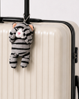 A gray tabby cat plush charm with its paws covering its eyes, hanging from the handle of a cream-colored suitcase by a silver ball chain.