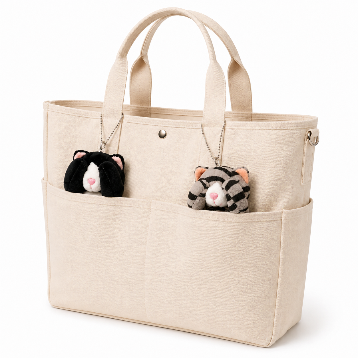Two cat plush charms, one gray tabby and one tuxedo cat, attached to the handles of a cream-colored tote bag with silver ball chains and tucked into the outer pockets.
