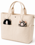 Two cat plush charms, one gray tabby and one tuxedo cat, attached to the handles of a cream-colored tote bag with silver ball chains and tucked into the outer pockets.
