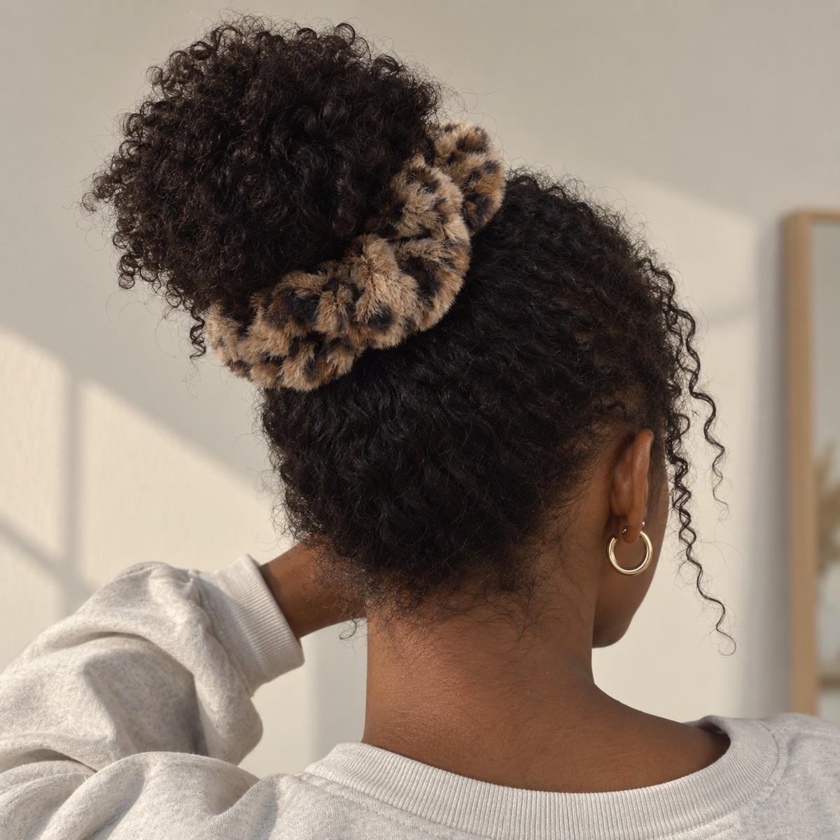 Woman wearing a soft, fluffy leopard print scrunchie with cat ears in beige, brown, and black to style her hair in a bun on top of her head.