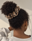 Woman wearing a soft, fluffy leopard print scrunchie with cat ears in beige, brown, and black to style her hair in a bun on top of her head.