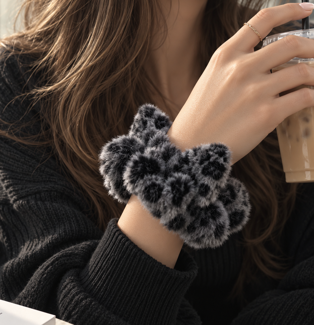 A close-up of a person wearing a fluffy leopard-print hair scrunchie in black and gray, holding an iced coffee. The person is wearing a black ribbed knit sweater.