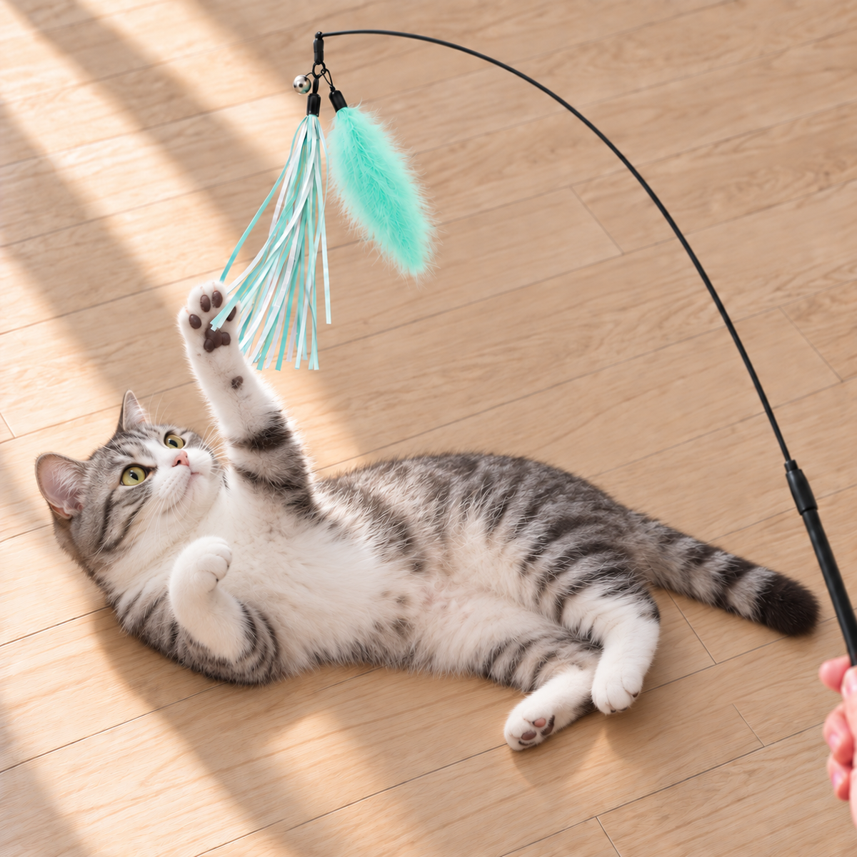 Gray and white cat lying on a wooden floor while playing with a mint green feather and ribbon cat wand toy.