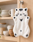 White cat-shaped hand towel with black embroidered details hanging from a hook on a wooden kitchen cabinet door.
